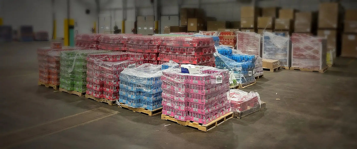 Stacks of colorful, plastic-wrapped beverage cans in red, green, blue, and pink hues are neatly arranged on wooden pallets within a warehouse.